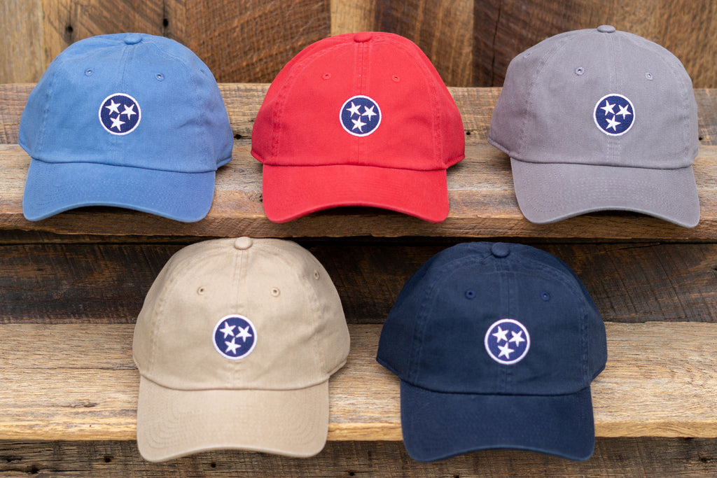 Volunteer Traditions Tennessee Tristar Hats. Stone Blue with Navy, Red with Navy, Old South Grey with Navy, Khaki with Navy, and Navy with Navy Tristar Hats on wood. 